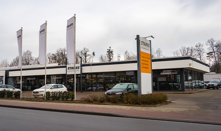 Auf dem Bild ist der Gebrauchtwagen Standort von STARKE Georgsmarienhütte an der Niedersachsenstraße zu sehen. Im Vordergrund wehen 3 Fahnen, das lange einstöckige Gebäude hat vor dem Eingang den Schriftzug "STARKE". Der Himmel ist grau und die Bäume blätterlos. 