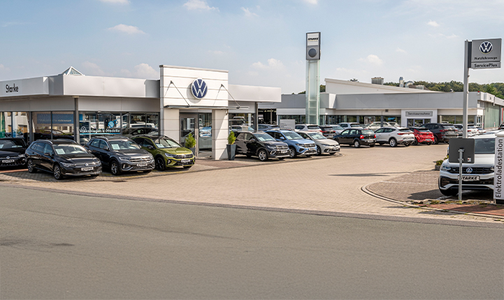 Auf dem Bild ist der Volkswagen Standort in Georgsmarienhütte zu sehen. Im Vordergrund steht der Eingang mit einem Volkswagen Logo. Im Hintergrund ist der geräumige Parkplatz zu sehen. 