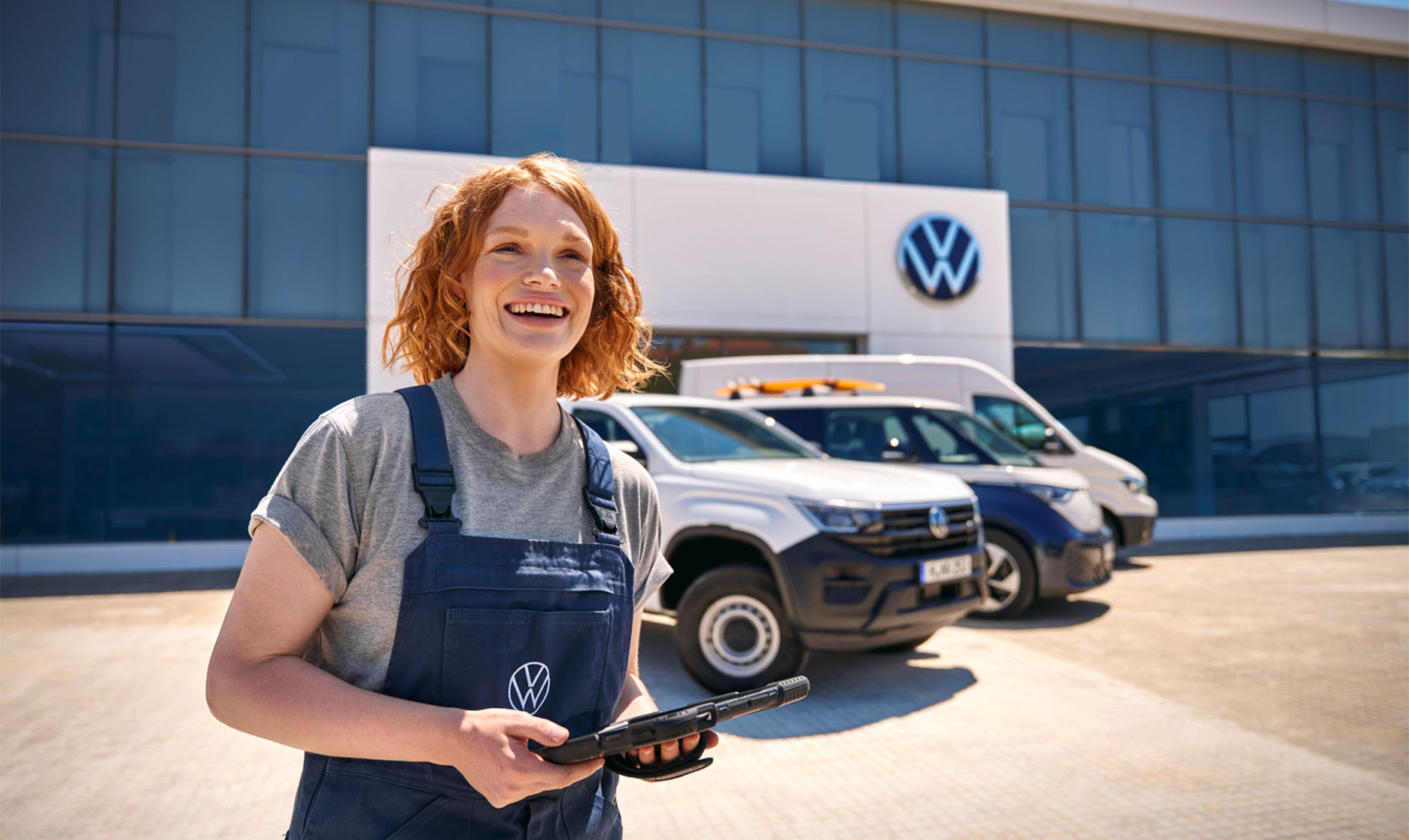 Im Vordergrund steht eine freundliche Frau in einem blauen Werkstatt-Overall von Volkswagen. Hinter ihr sind einige VW Nutzfahrzeuge vor einem VW Autohaus aufgestellt. 