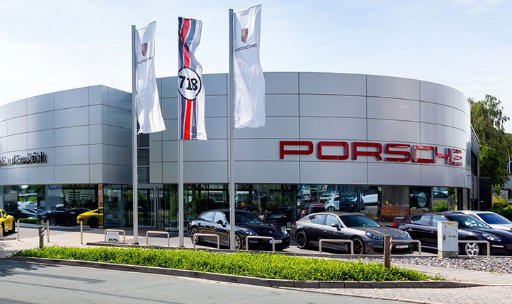 Auf dem Bild ist das Porsche Zentrum der STARKE Automobilgruppe am Blumenhaller Weg abgebildet. Die Bäume im Hintergrund sind grün und im Vordergrund wehen Porsche Fahnen. 