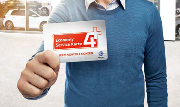 Das Bild zeigt den Oberkörper von einem Mann der einen blauen Pullover mit einem Hemd darunter trägt. In seiner rechten Hand hält der eine Karte auf der Economy Service Karte 4+ jetzt Vorteile sichern steht. Im Hintergrund ist eine Gebäude in den Fahrzeuge stehen zu sehen.