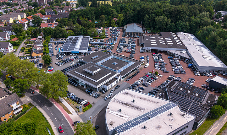 Luftaufnahme VW Zentrum mit mehreren Ausstellungsflächen, Fahrzeugstellplätzen und Gebäuden in einer von Bäumen umgebenen Umgebung.