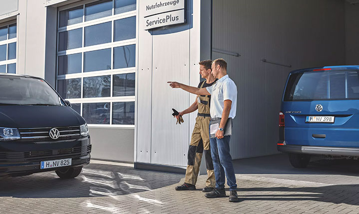 Das Bild zeigt einen Serviceberater und einen Mechaniker vor eine VW Werkstatt. An der Werkstatt ist das Schild Nutzfahrzeuge Service Plus angebracht. Rechts im Bild steht ein blauer Multivan der nur von hinten zu sehen ist in der Werkstatt. Neben der Werkstatt steht ein schwarzer Multivan der nur von vorne zu sehen ist.